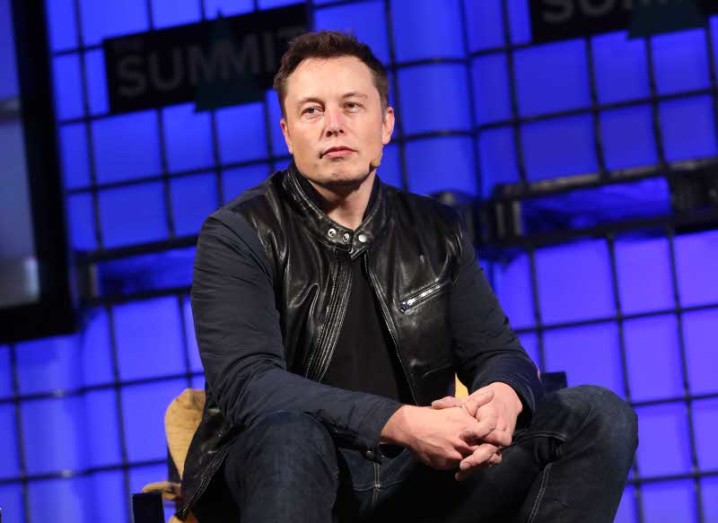 Elon Musk is dressed all in black, sitting on a stage with blue lights behind him.