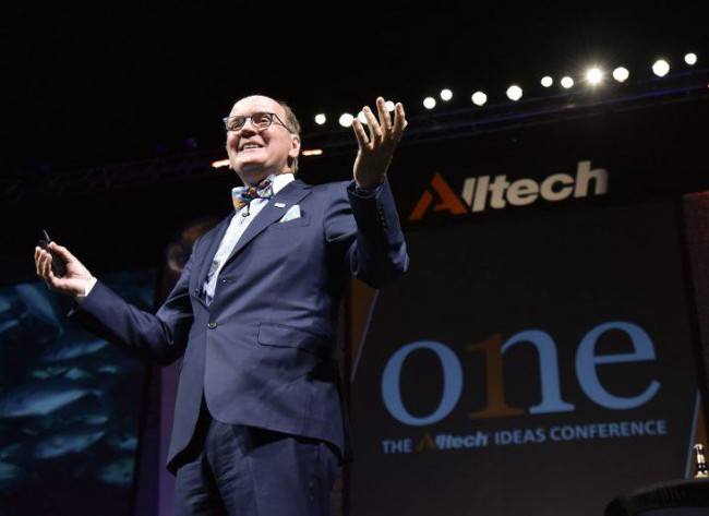 Man in dark suit and dickie-bow stands on a stage with his arms open wide.