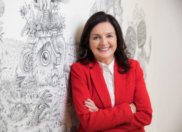 woman with black hair wearing bright red blazer and white shirt smiling broadly with arms folded against a white wall with grey pattern.