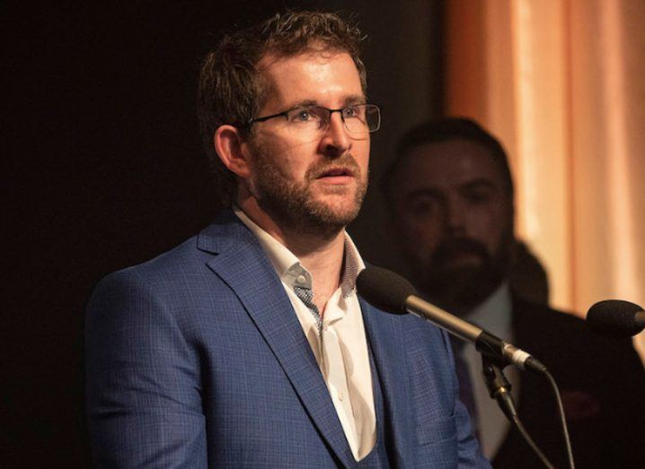 man in glasses and navy suit speaking into mic while standing at podium.