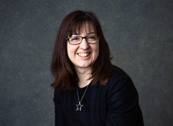 A headshot of Catherine Breslin dressed in black and standing against a dark grey background. She’s smiling at the camera.