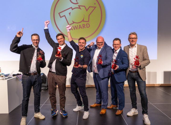 A group of six men stand together cheering while holding red awards at a toy award show.