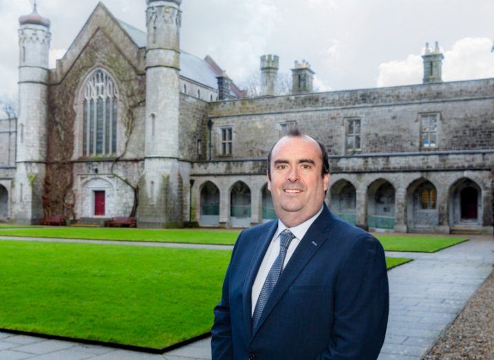 Prof Peter Doran standing outside the stone Quadrangle building at University of Galway with a patch of green grass behind him in front of the building.
