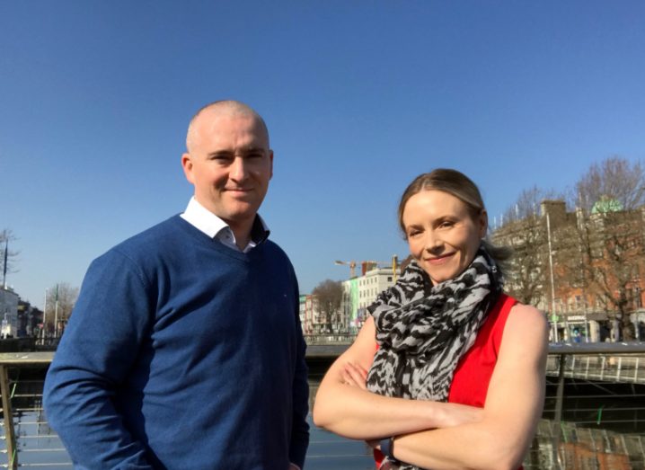 Legitimate founders Gerard and Caoimhe Donnelly stand on the Rosie Hackett Bridge in Dublin city centre on a clear sunny day. The River Liffey leading towards O’Connell Bridge is visible in the background.
