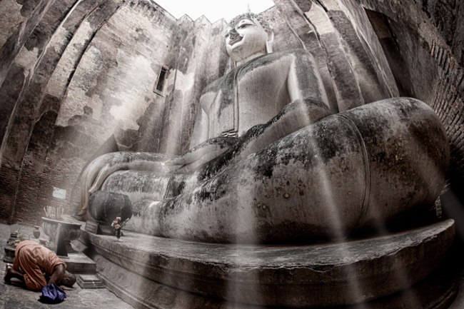 A monk paying homage to the Buddha statue of Wat Si Chum, Sukhothai Historical Park, Thailand. Image via Yakuzakorat/Wikimedia Commons