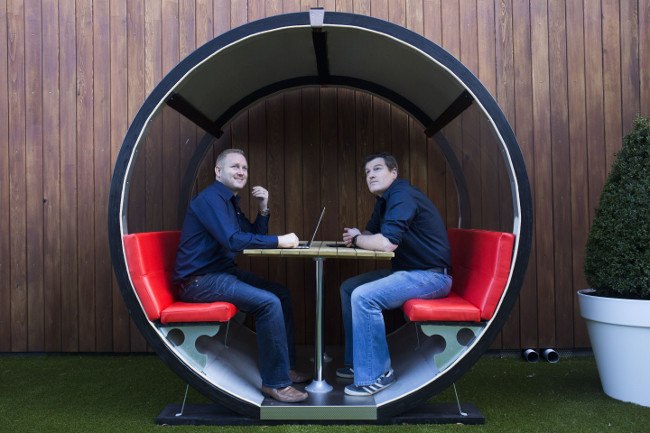 Two men in blue shirts and jeans sit inside a circular seating unit with red leather seats.