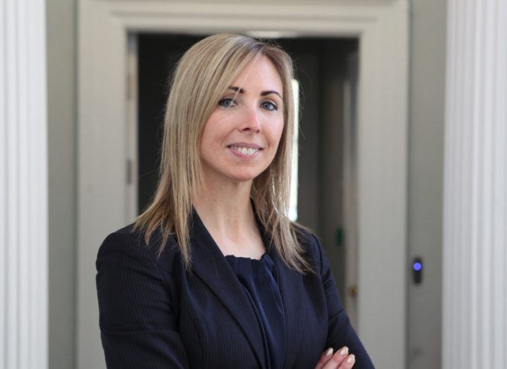 Blonde woman in dark suit with arms folded, smiling.