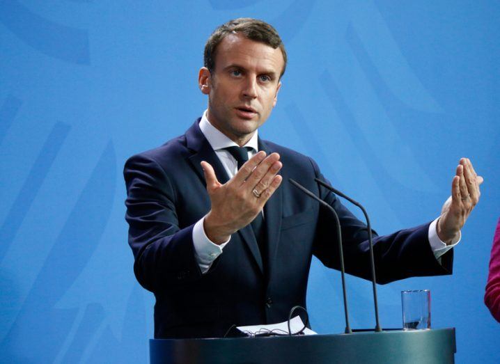 Emmanuel Macron stands at a podium in a suit, speaking to a crowd.