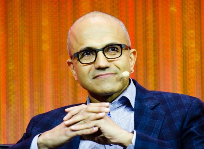 Satya Nadella sits on a stage wearing a microphone up to his face.