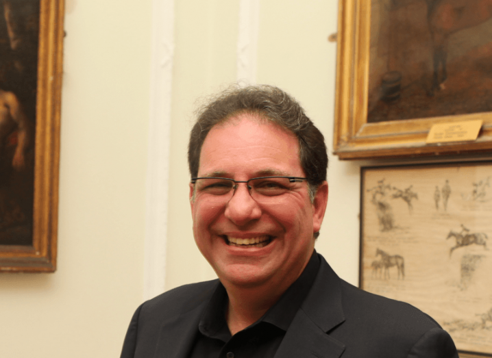 Kevin Mitnick smiling at the camera in a dark suit with portraits in the wall behind him.