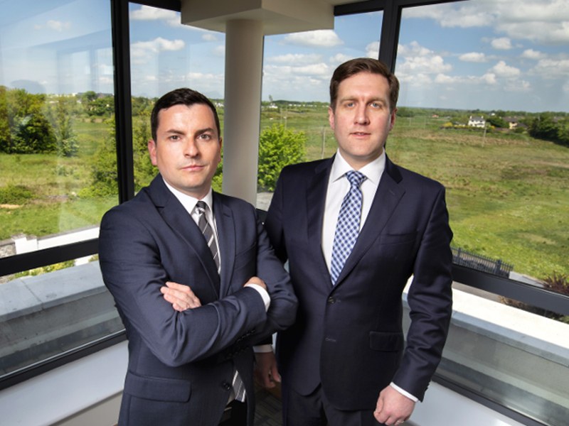 Two men in suits with a green field and blue sky in the background.