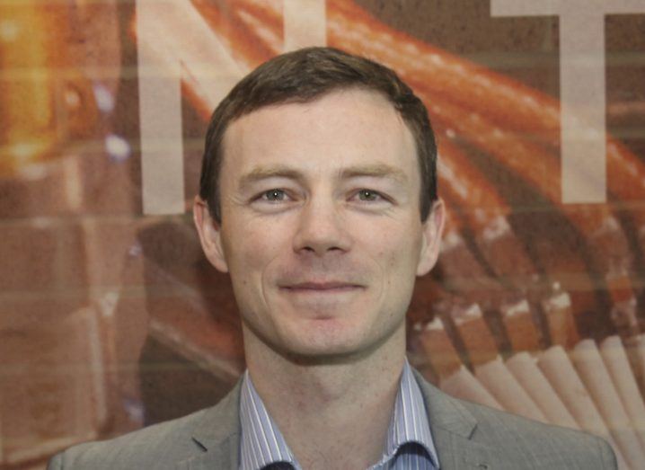 Headshot of a man in a suit, smiling at the camera in front of a brick wall featuring artwork.