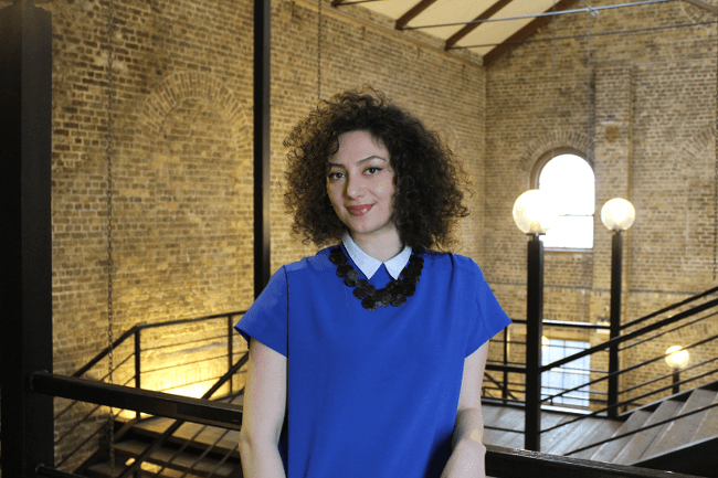A woman with short, dark curly hair in a royal blue shirt dress poses by a rail overlooking a spacious stairway surrounded by brickwork walls