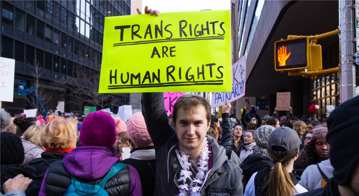 A man wearing a lei garland stands amid a crowd of women in New York holding a brightly coloured protest sign that reads ‘trans rights are human rights’