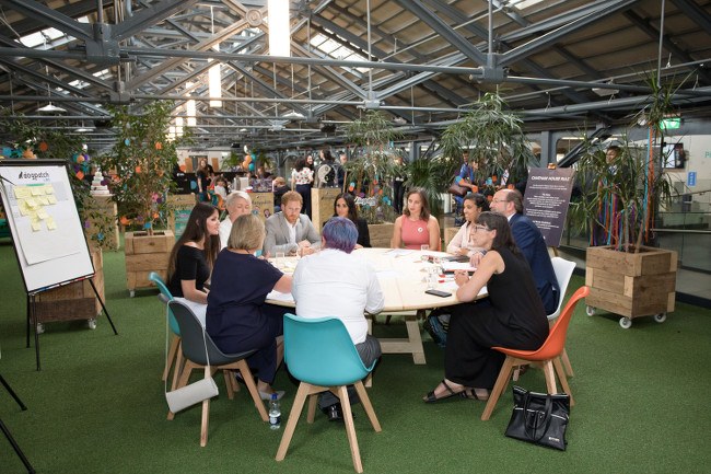 Participants seated at a round table with Prince Harry and Meghan Markle in an open space in Dogpatch Labs at the CHQ building