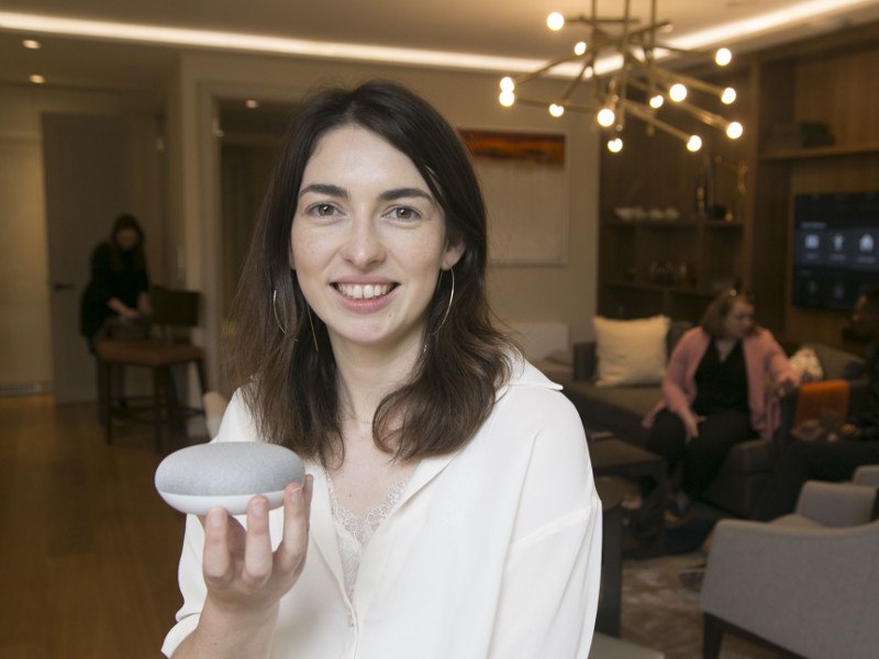 A woman with dark hair and a white blouse holds a Google Home Mini in her hand.
