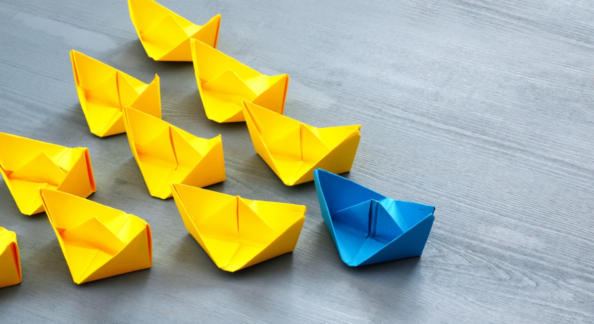 A blue paper sailboat leading a bunch of yellow paper sailboats on a wooden surface to represent leadership skills.