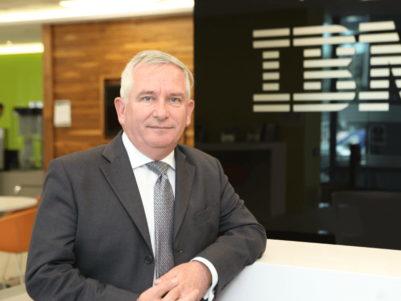 Paul Farrell smiling wearing a suit with the IBM logo in the background.