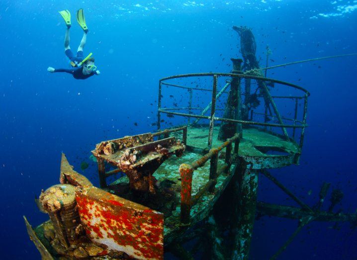 The tower of a submerged, rusted shipwreck with a diver approaching it.