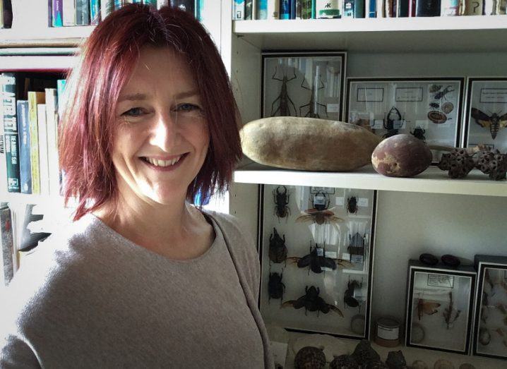 Colette Kinsella stands smiling in front of a bookcase packed with books, rocks, shells and insect displays