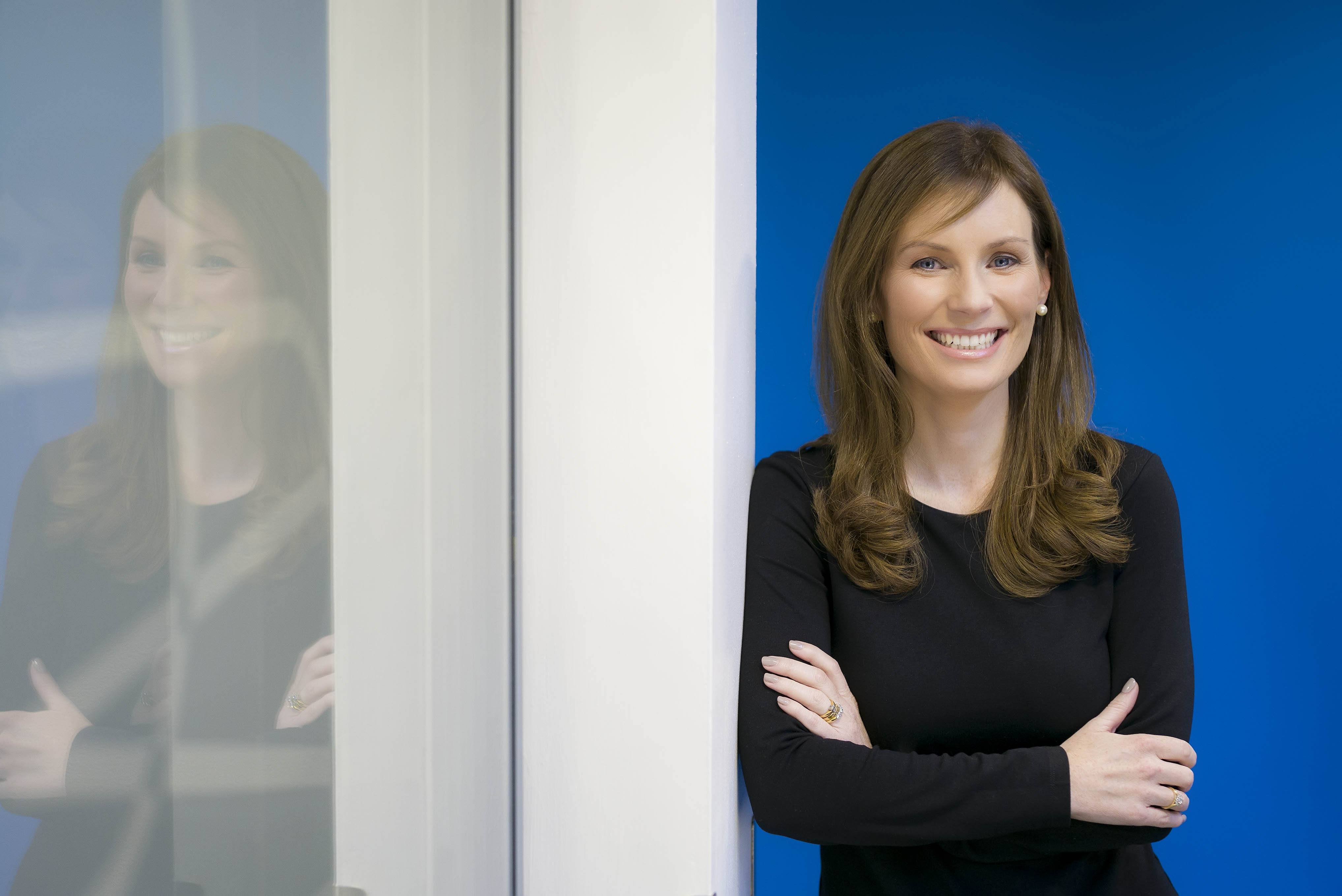 young woman with long brown hair in black top with arms folded leaning against wall with reflection in nearby window.