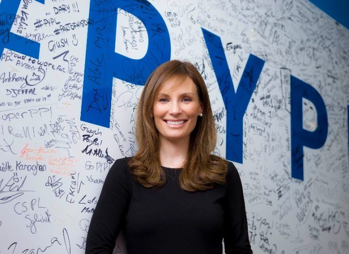 young woman with long brown hair in black top standing in front of white wall covered in signatures