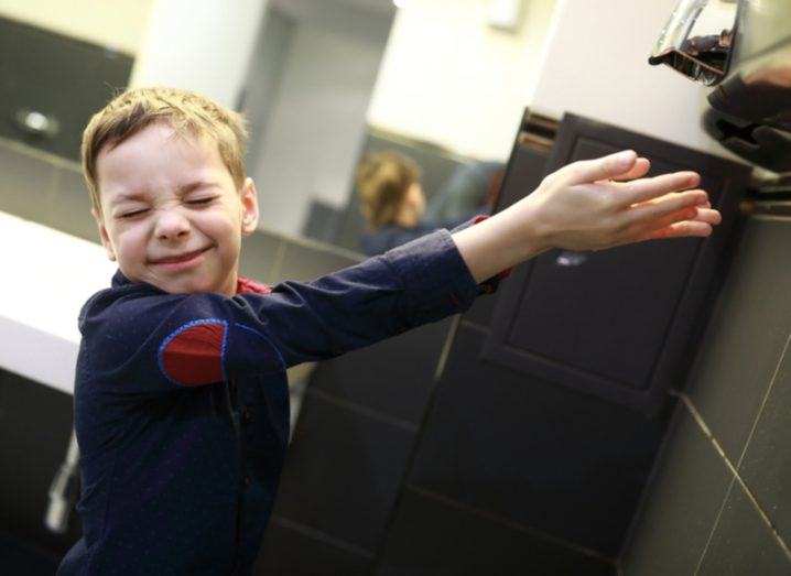 A grimacing child holding his hands out under an air hand dryer.