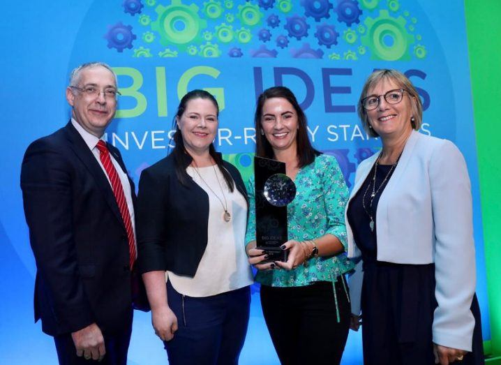 A man and three women, one of whom is holding a trophy, stand in front of a blue and green Big Ideas backdrop.