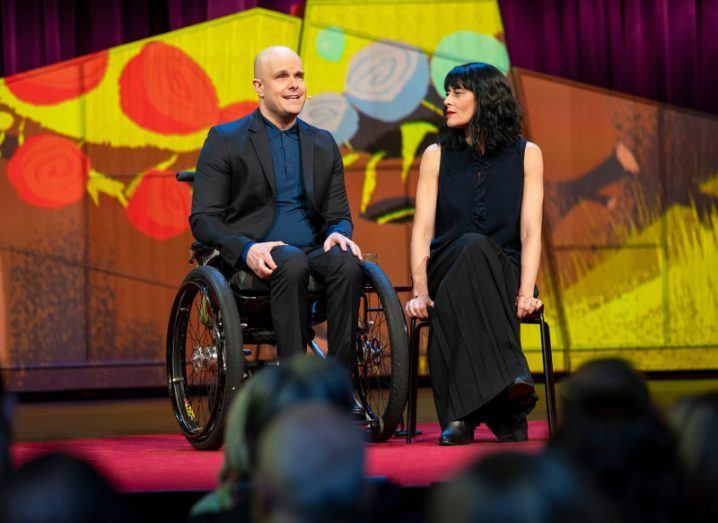 A bald man in a wheelchair speaks to an audience from a colourful stage, while a dark-haired woman seated next to him looks on.