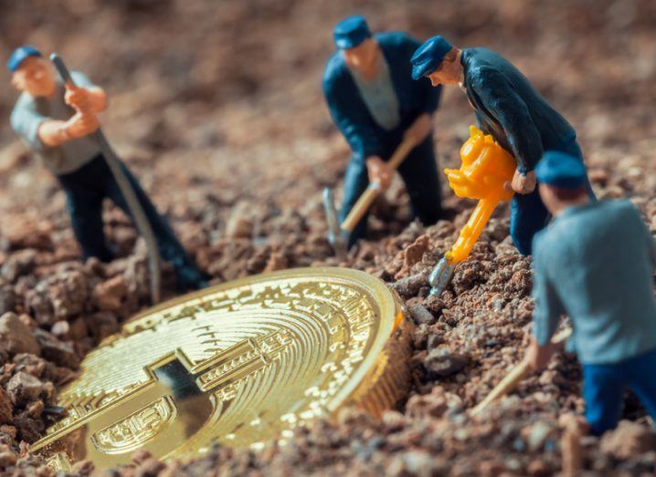 toy figures representing miners dig a cryptocurrency coin out of the sand.