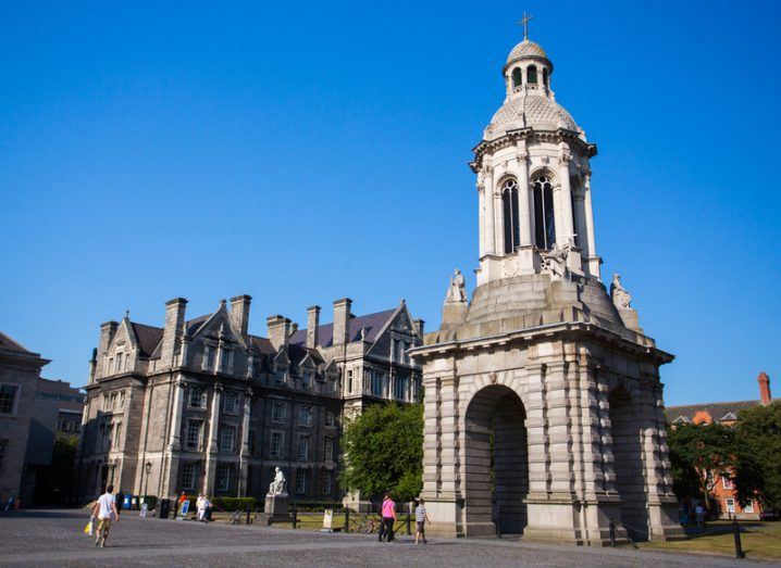 People walking across Trinity campus in Dublin beside old buildings under a blue sky.