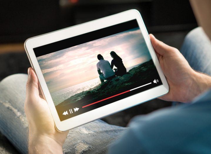 Over-the-shoulder view of a man watching video of a couple on the beach on his tablet.