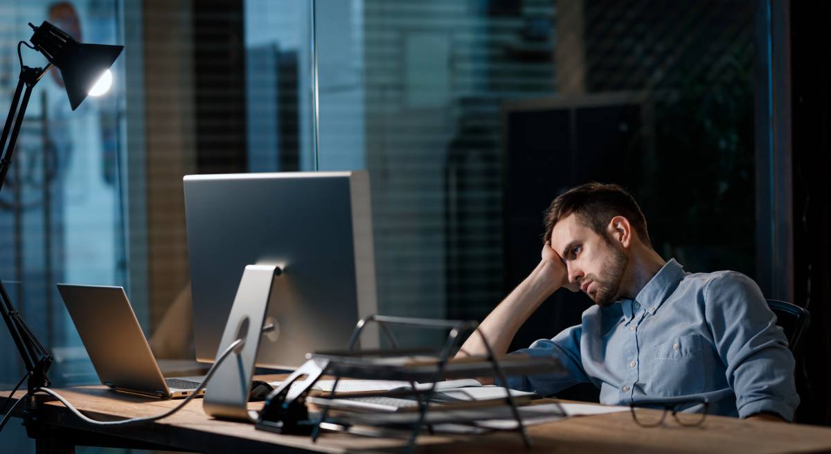 Casual man looking fatigued while working with computer in dark office alone.