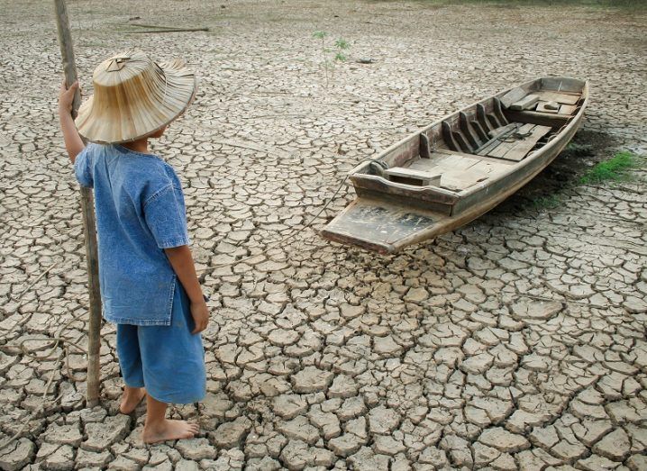 A boy in blue overalls standing on a cracked, dried out riverbed looking at a small boat.