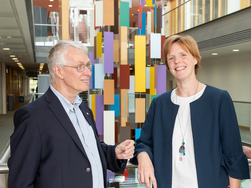 In front of a colourful hanging sculpture, a grey-haired man smiles at a woman with short hair who is looking at the camera.