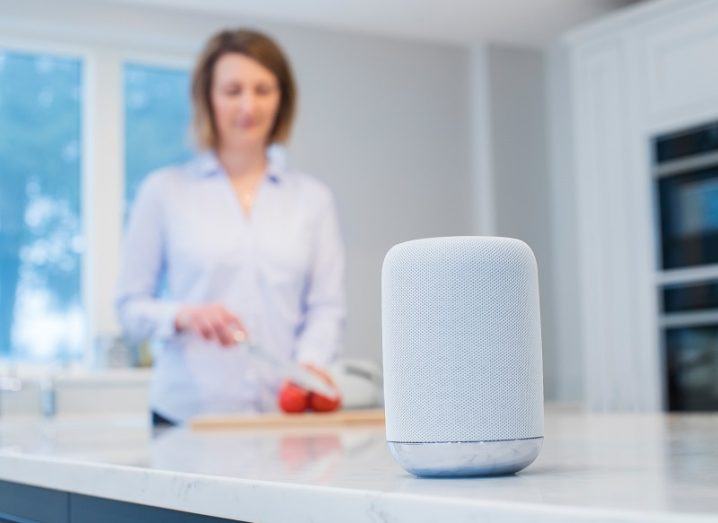 Woman chopping tomatoes in the kitchen with a smart speaker in the foreground, listening.