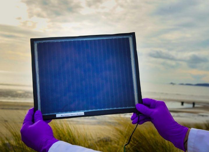 A scientist with purple gloves holding an A4 sheet-sized solar cells up in the air.