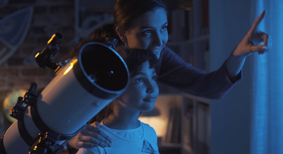 A woman with a young girl smiling and pointing out at the sky, standing beside a telescope dreaming about space careers.