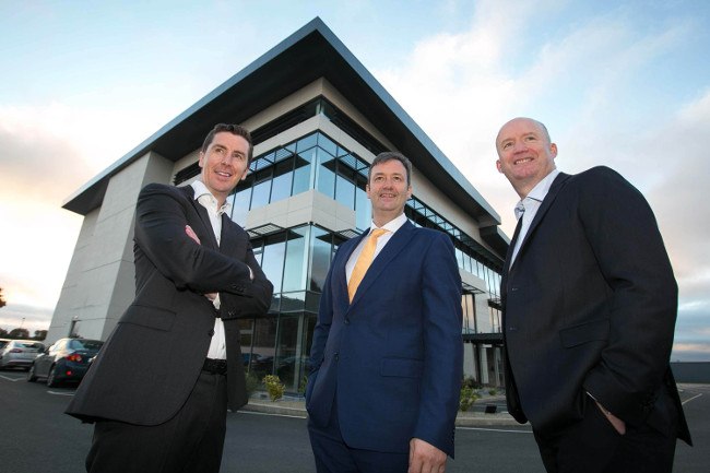 Three men in suits stand before a building under a cloudy blue sky.