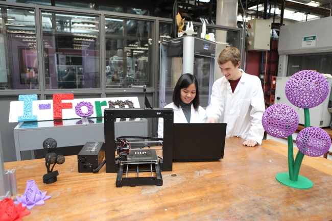 Wide view of two scientists working at a bench with a 3D printer and laptop, surrounded by colourful printed objects.