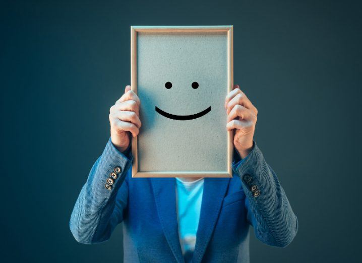 A person in a blazer holds a frame containing a smiley face in front of their head.