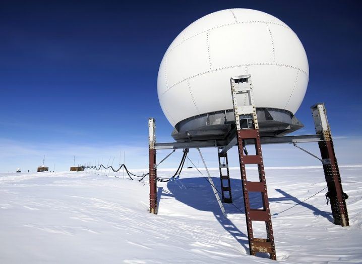 A white telecoms dome on four legs against the backdrop of Antarctic ice and blue sky.