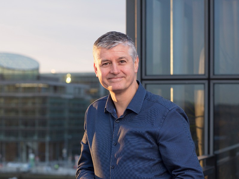 middle aged man with grey hair wearing dark blue shirt and smiling, standing outside on balcony, with setting sun and city landscape in background.
