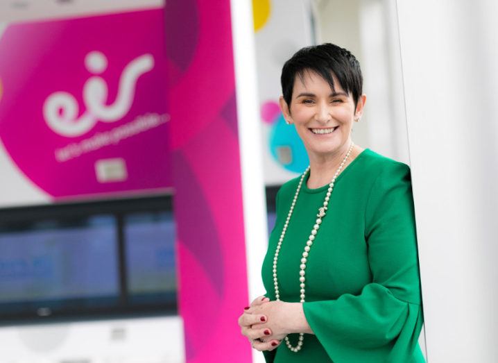 Woman in green dress with pearls smiles at camera with Eir sign in background.
