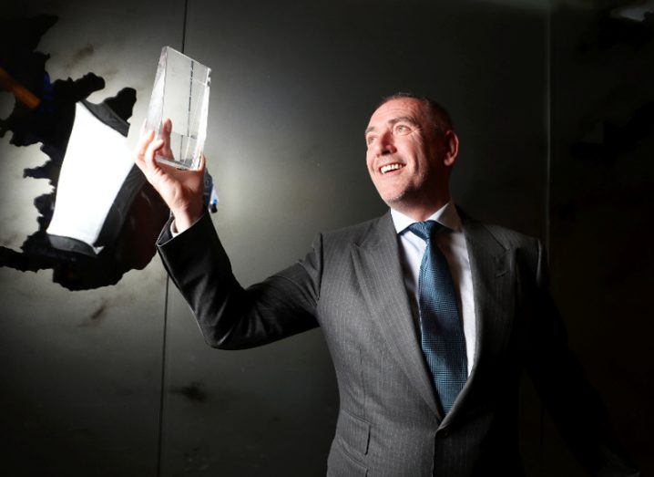 Man in grey suit holds up a crystal award.