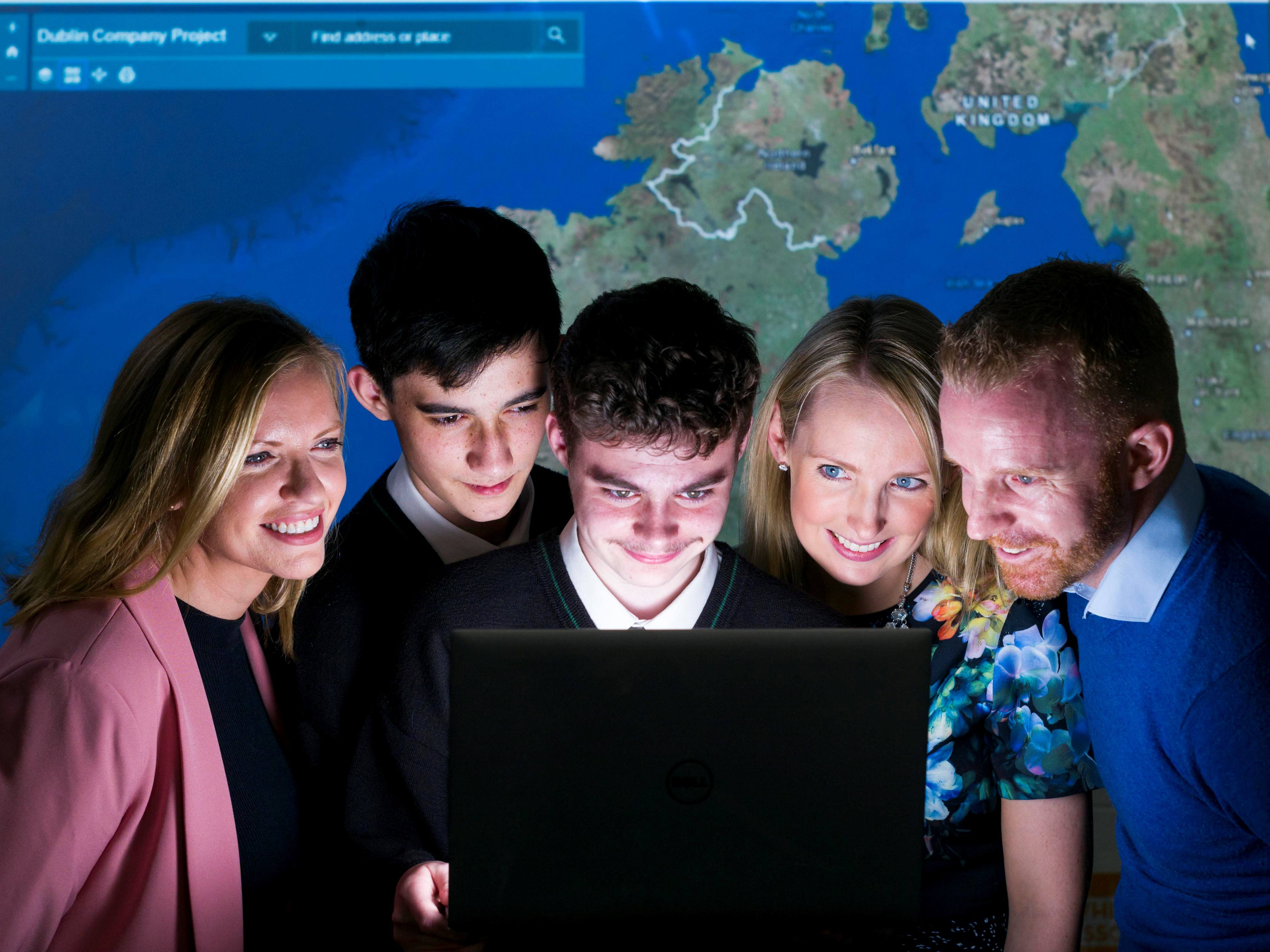 Two women, two boys and a male teacher look into a laptop screen with a map in background.