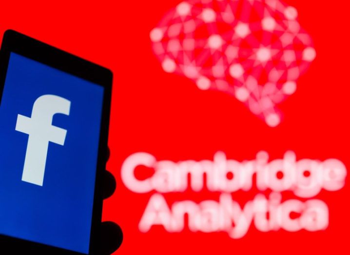 Smartphone in hand with logo of popular social network Facebook. Cambridge Analytica emblem in background.