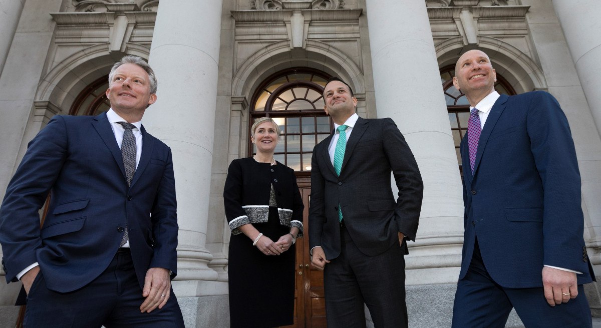 three men in dark suits and a woman in a dark dress stand outside government buildings in Dublin.