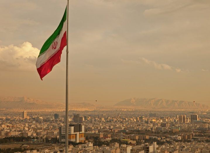 Iran flag waving in the wind above skyline of Tehran lit by the orange glow of sunset.