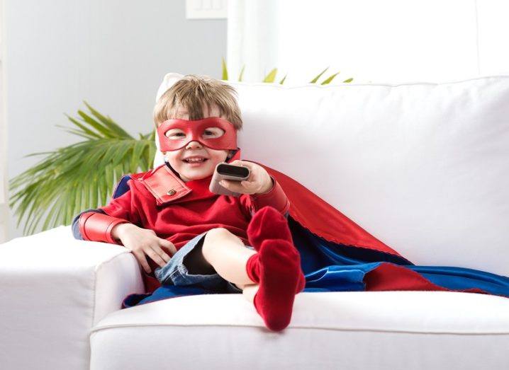 Smiling young boy in a superhero outfit sitting on a couch watching a movie.